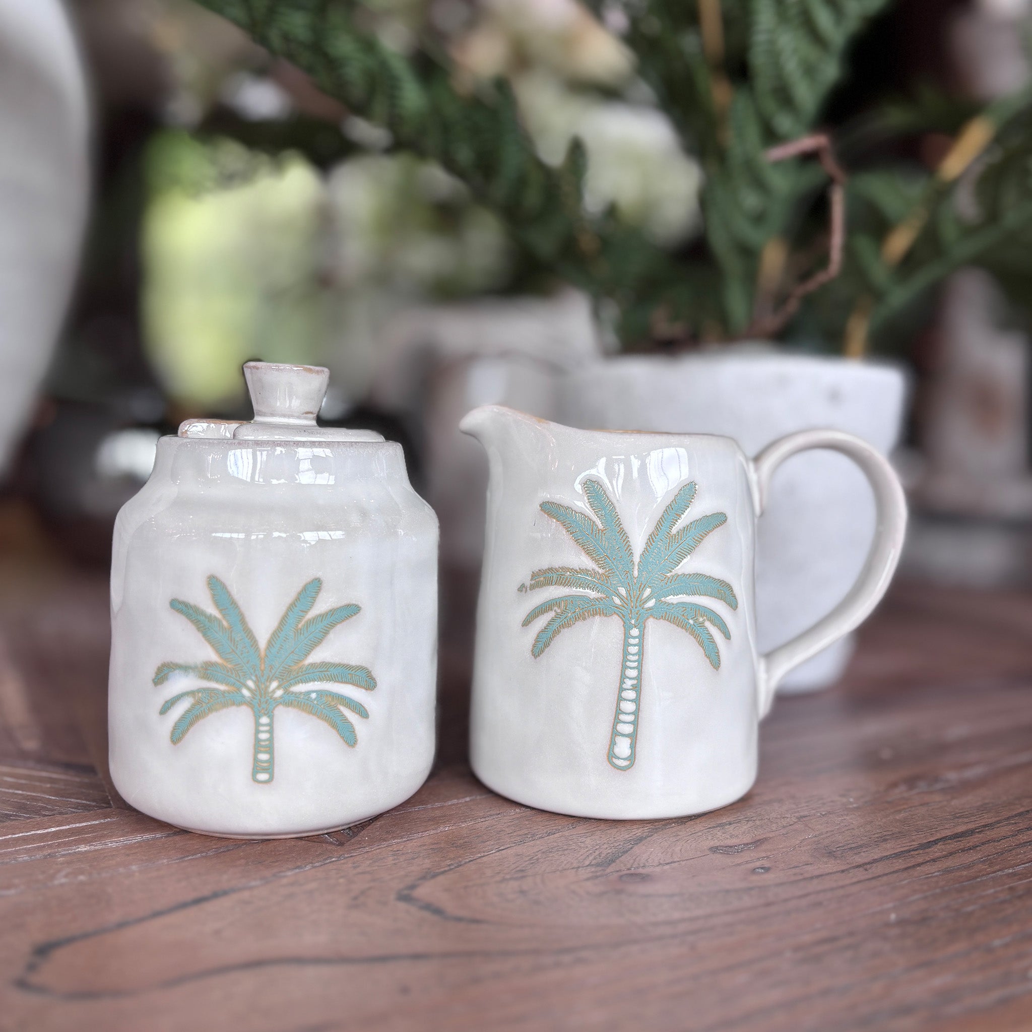 A charming ceramic milk jug and sugar pot set, finished in a soft white glaze with warm sandy tones and delicate gold-edged detailing, and a hand-painted style palm tree motif in muted greens and gold.