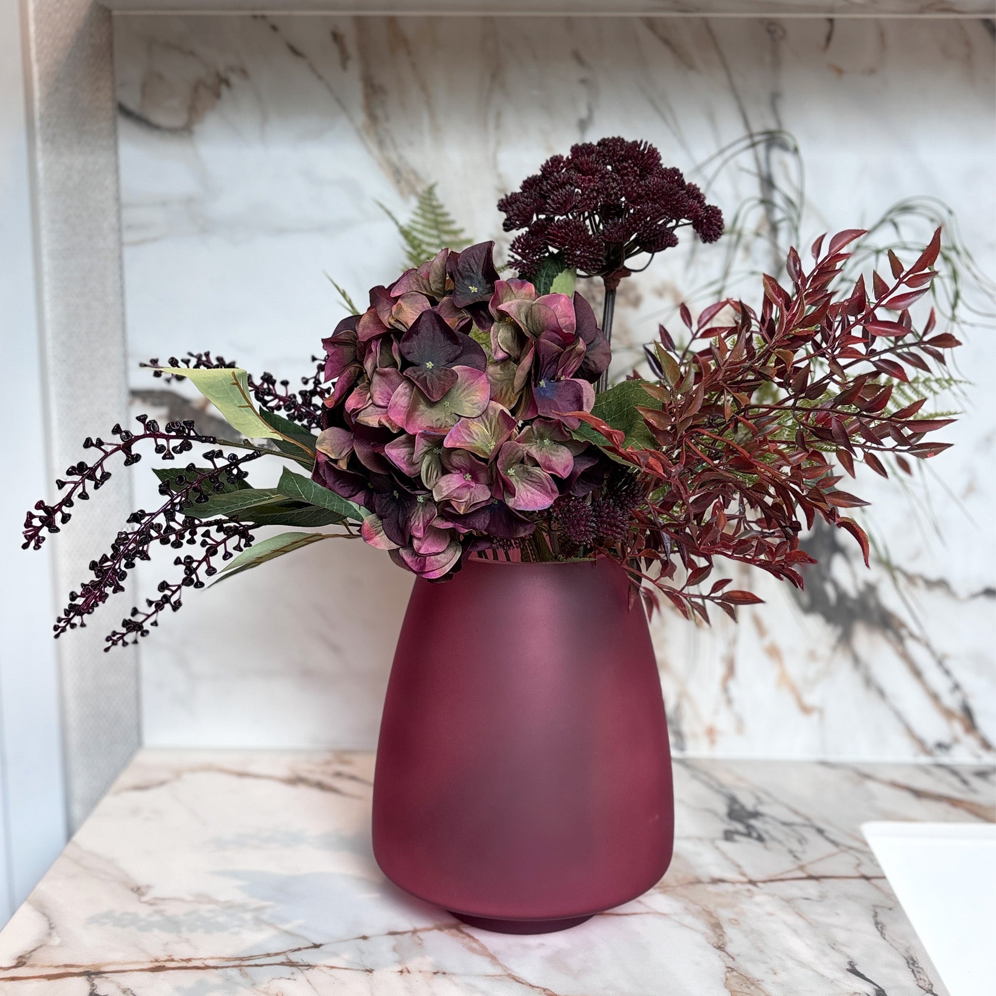 A rich, abundant display centred on plum hydrangeas and deep burgundy angelica seed heads, softened with delicate green fern and wispy grasses, and finished with dark berry sprays and russet foliage, in a claret red glass vase. Overall Height: 70cm, Overall Width: 60cm, Delivery is 2-10 working days, depending on stock availability