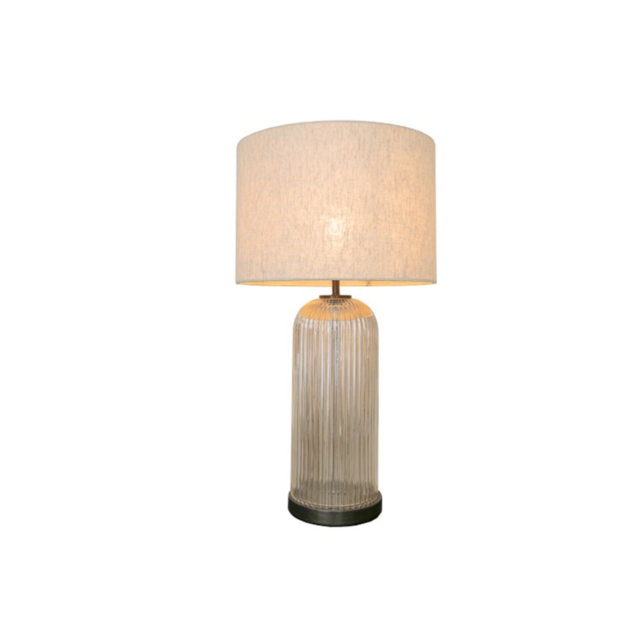 A elegant table lamp with a tall cylindrical ribbed glass base in a warm champagne tint, set on a dark metal base ring. Paired with a classic drum-shaped natural linen shade.